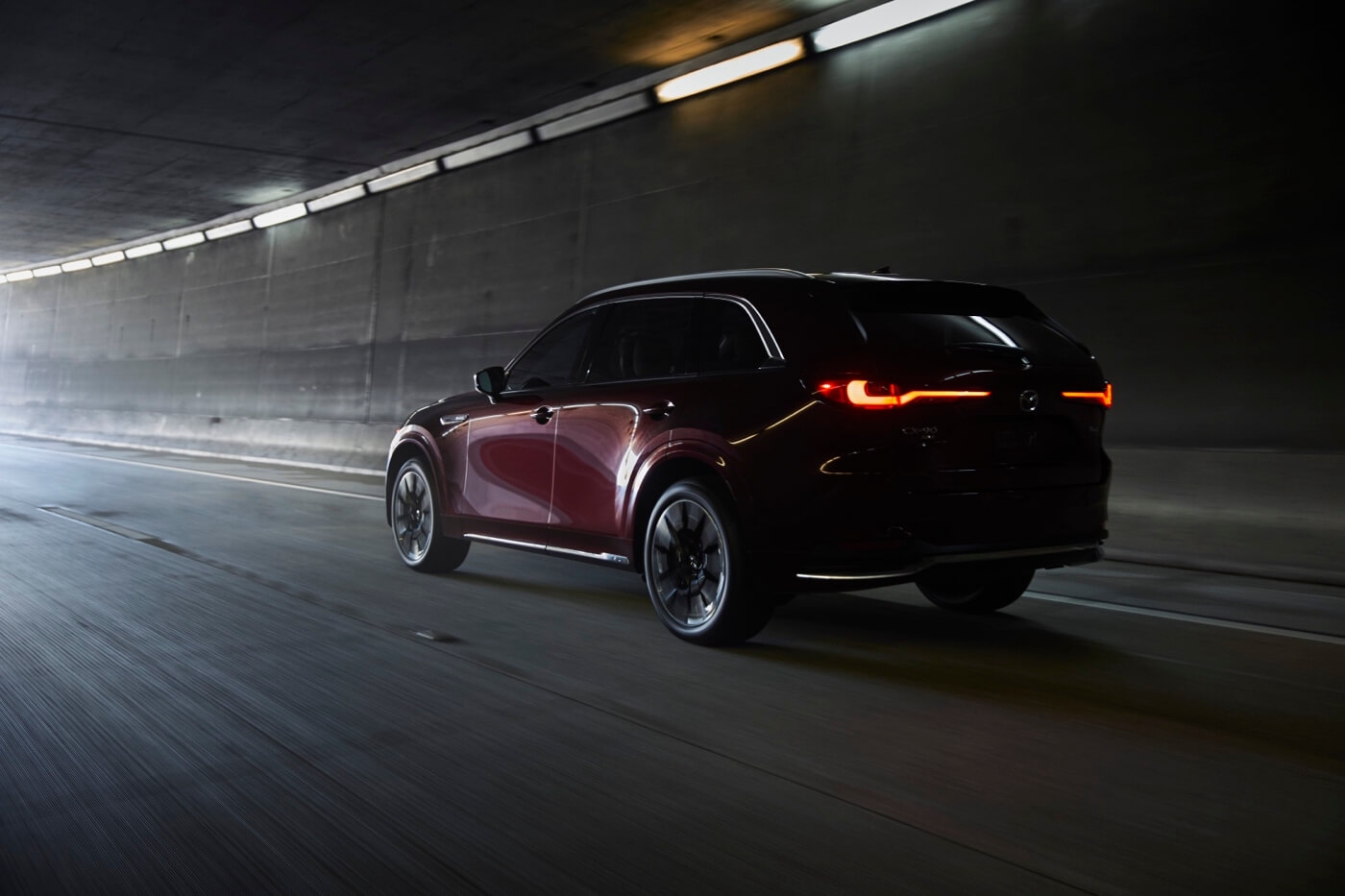 Mazda CX-90 PHEV 2024 rouge foncé vu de trois quarts arrière en mouvement dans un tunnel éclairé.